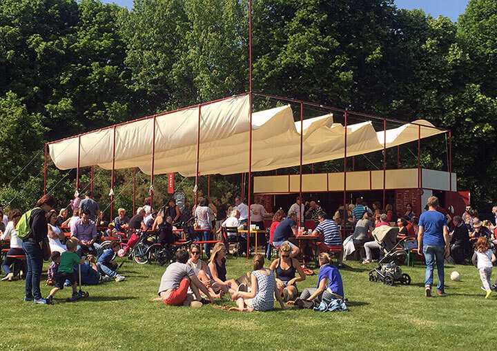Een park met een eettent, mensen zittend in het gras of aan tafel, en mensen op wandel