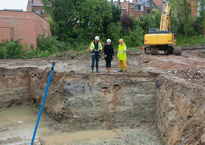 Drie personen met veiligheidshelmen kijken in een uitgegraven put met een kraan in de achtergrond