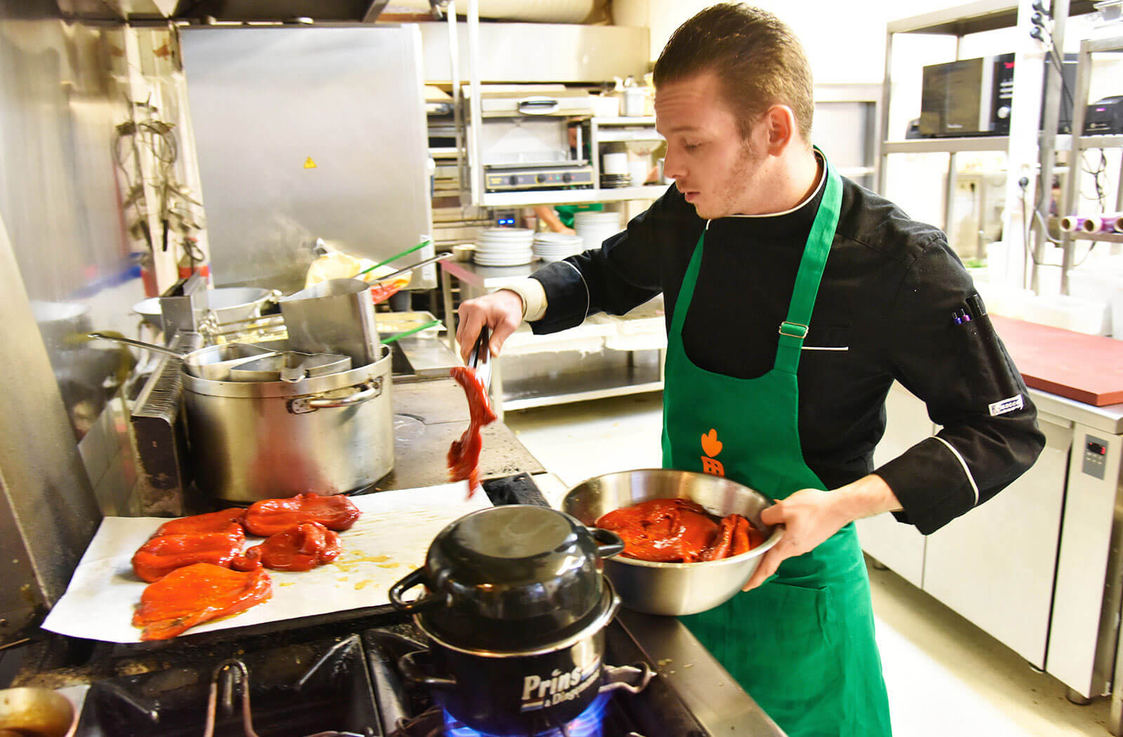 In een keuken van een restaurant legt een man stukken vlees in een pan. Hij draagt een groene schort met een oranje wortel erop.
