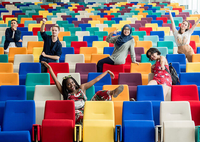 Zes personen die op een grappige manier poseren in een auditorium, op veelkleurige stoelen
